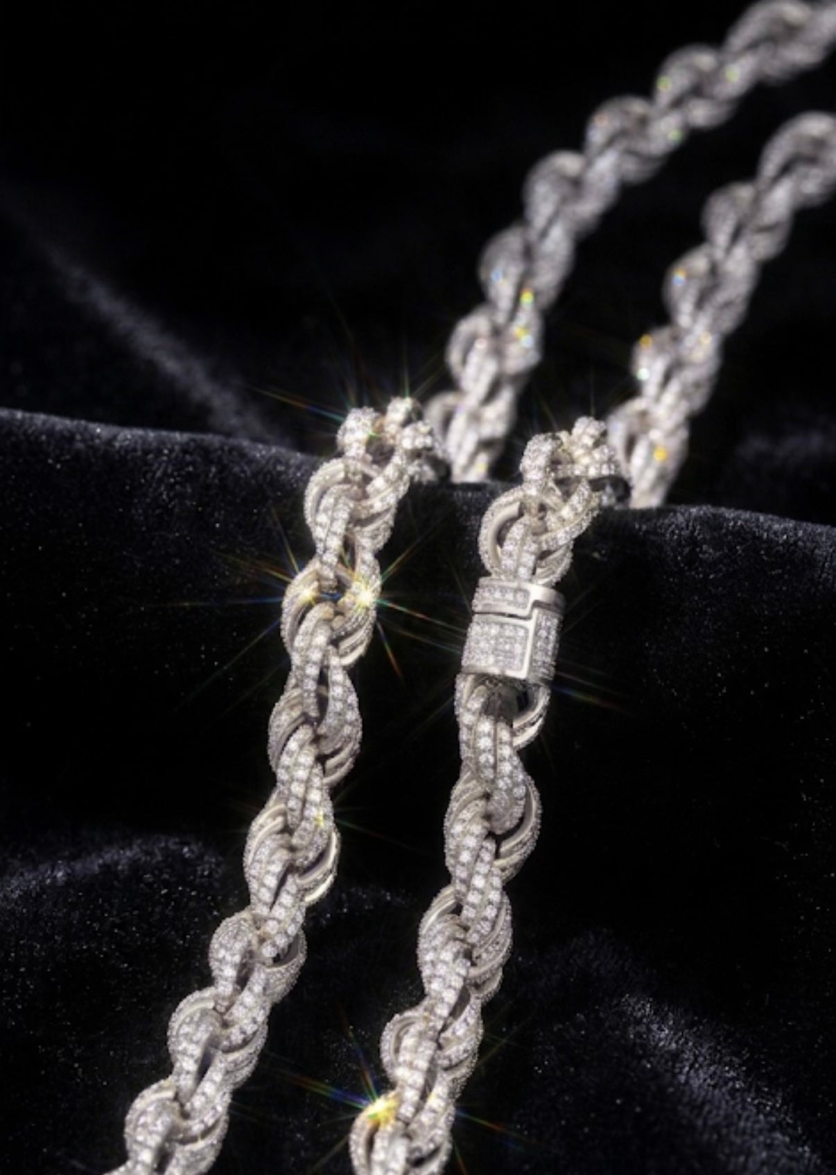 A close-up of a braided rope-style chain necklace with a rectangular clasp, covered in sparkling clear stones, displayed on a black velvet surface.