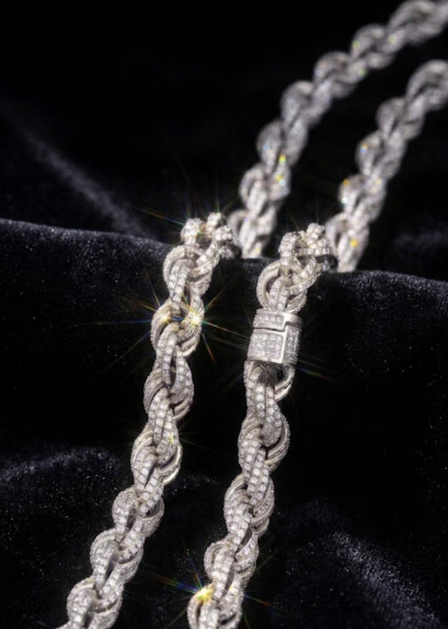A close-up of a braided rope-style chain necklace with a rectangular clasp, covered in sparkling clear stones, displayed on a black velvet surface.